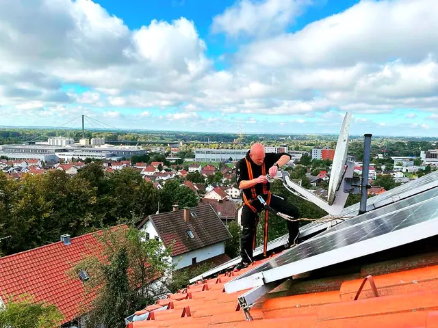 Monteur richtet auf einem Ziegeldach eine Sat-Schüssel aus, daneben liegen Photovoltaikmodule
