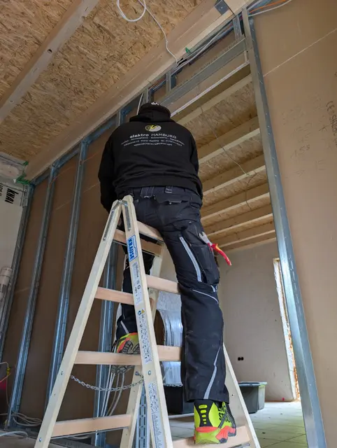 Monteur auf der Leiter verlegt Leitungen entlang der Dachschräge im Rohbau