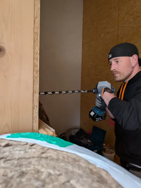 Monteur bohrt mit einem langen Holzbohrer einen Durchbruch für die Leitungsführung